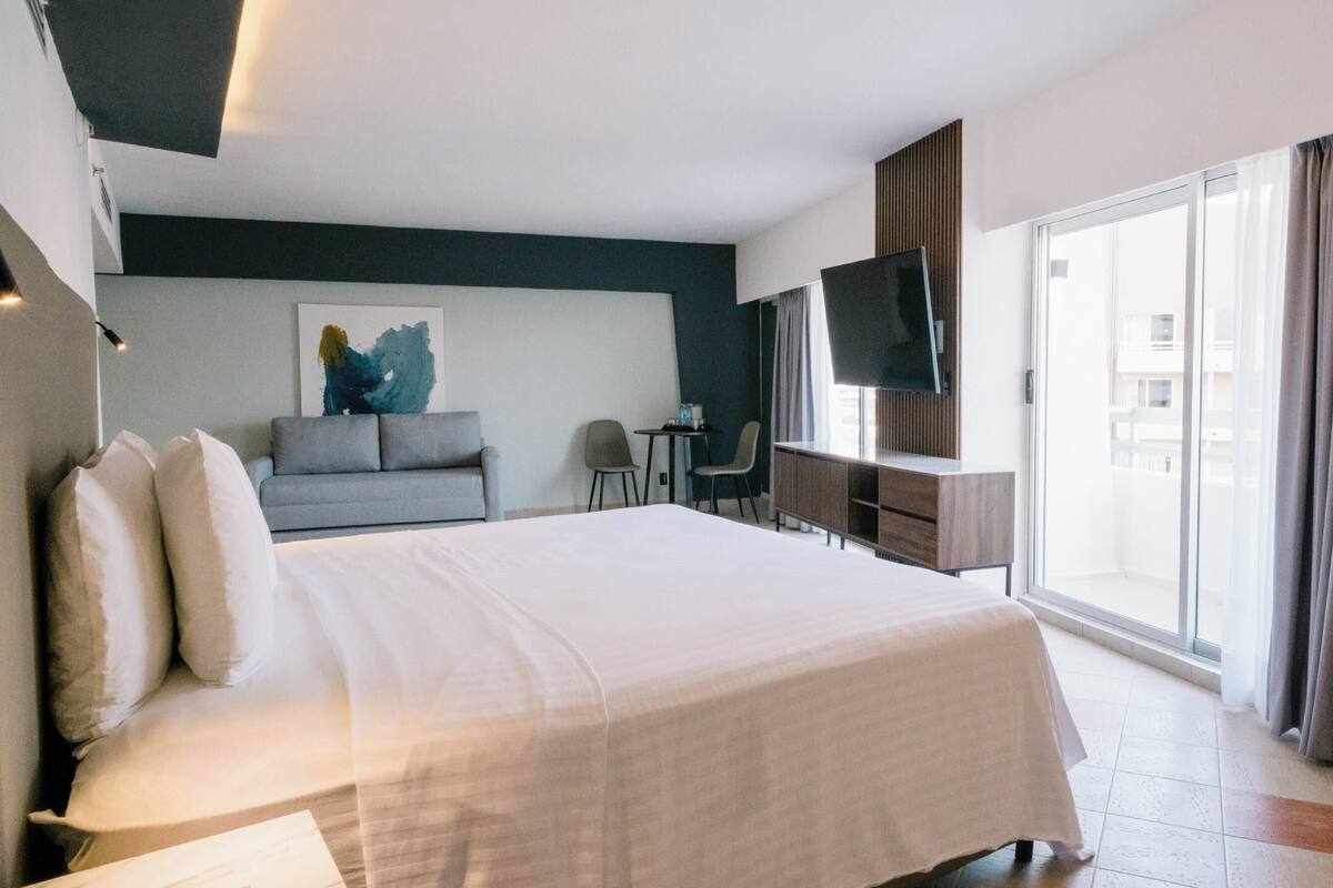 A hotel room with a large bed, a desk, a chair, a flat-screen TV, and a large window with curtains.