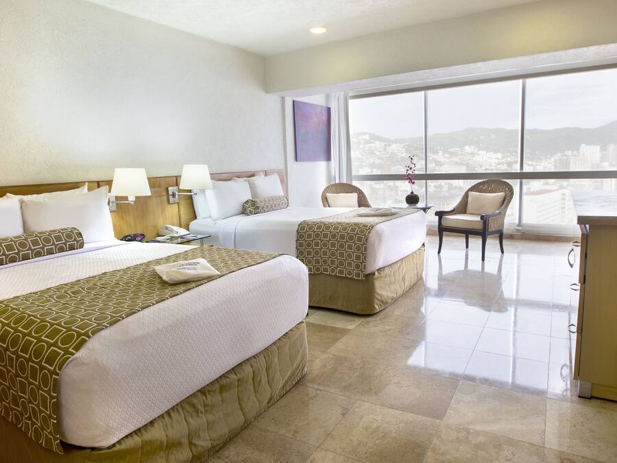 Habitación de hotel con dos camas, un escritorio, una silla, un televisor y un ventanal con vistas a la ciudad.