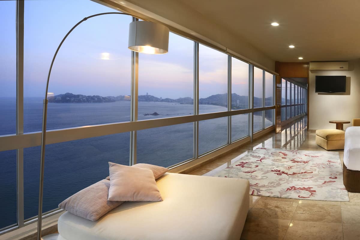 A hotel room with a large window offering a view of the sea and hills, a bed with a wooden headboard, a bedside table with a lamp, and a small seating area with a chair and ottoman.