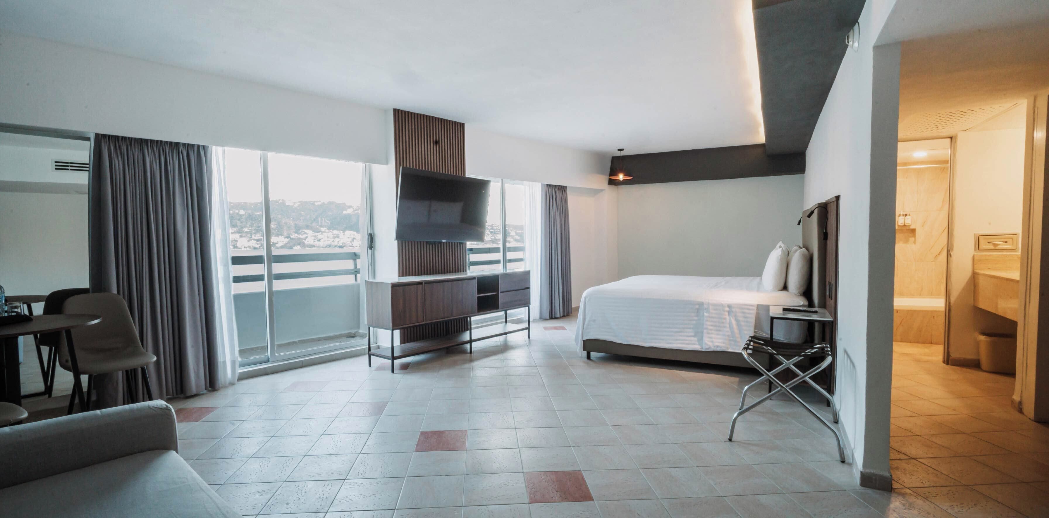 A hotel room with a bed, a TV, a desk, a chair, a sofa, a balcony with curtains, and a view of the city.