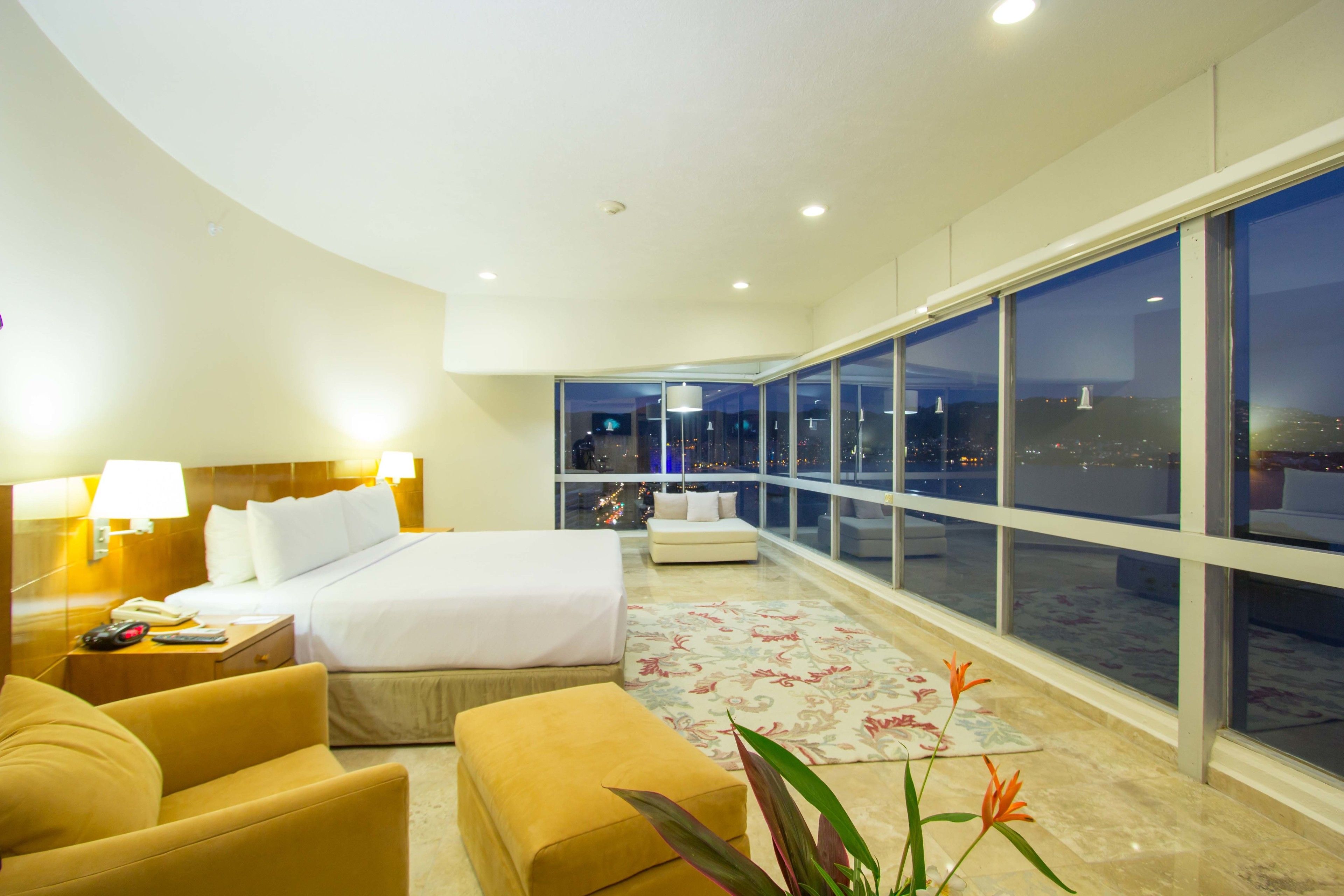 A modern hotel room with a large bed, a sofa, a desk with a lamp, and a view of the city at night.