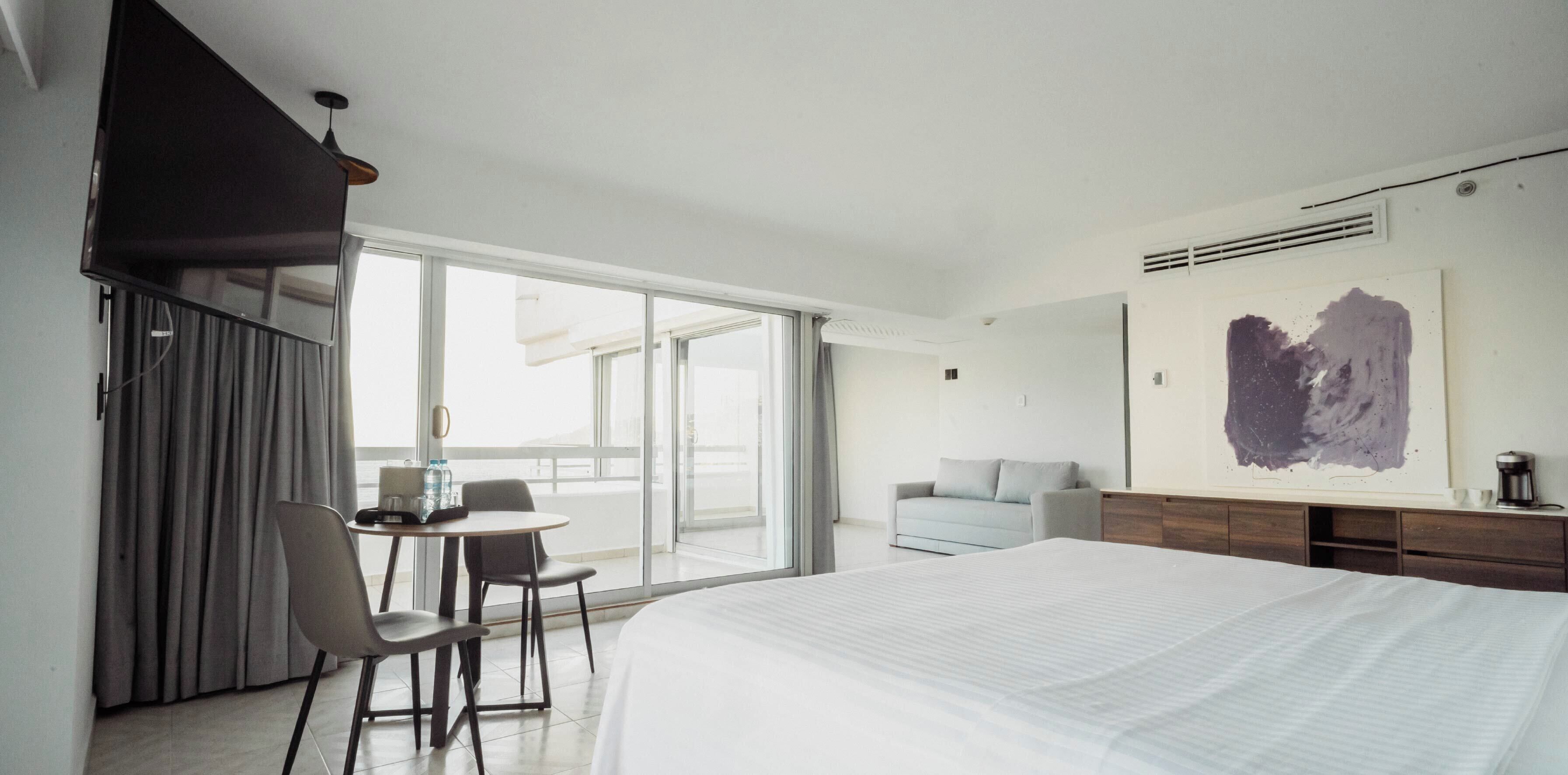 A modern hotel room with a large bed, a desk with chairs, a sofa, and a large window offering a view.