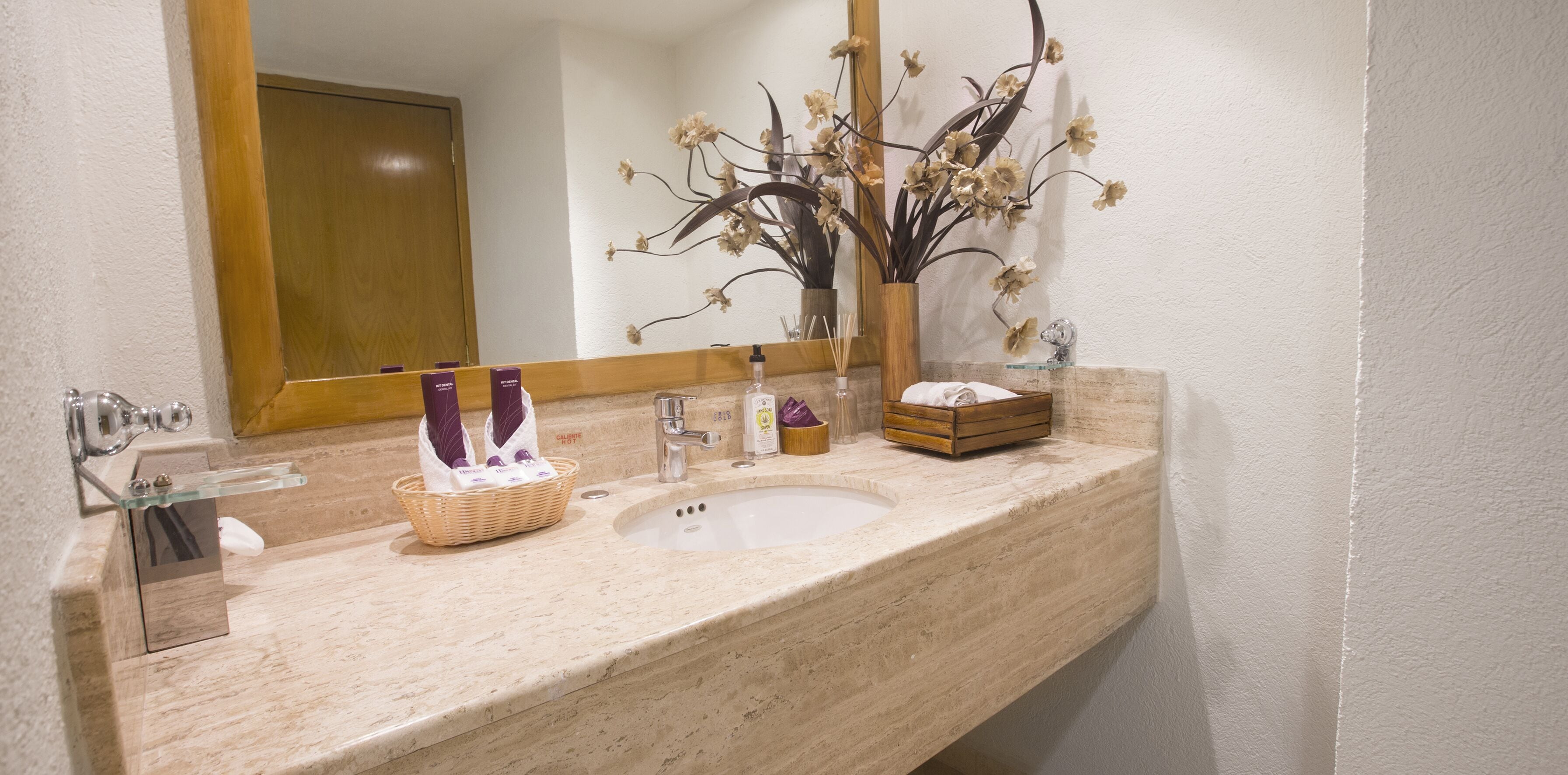 A bathroom with a marble sink, a mirror, a vase with dried flowers, and a basket with toiletries.
