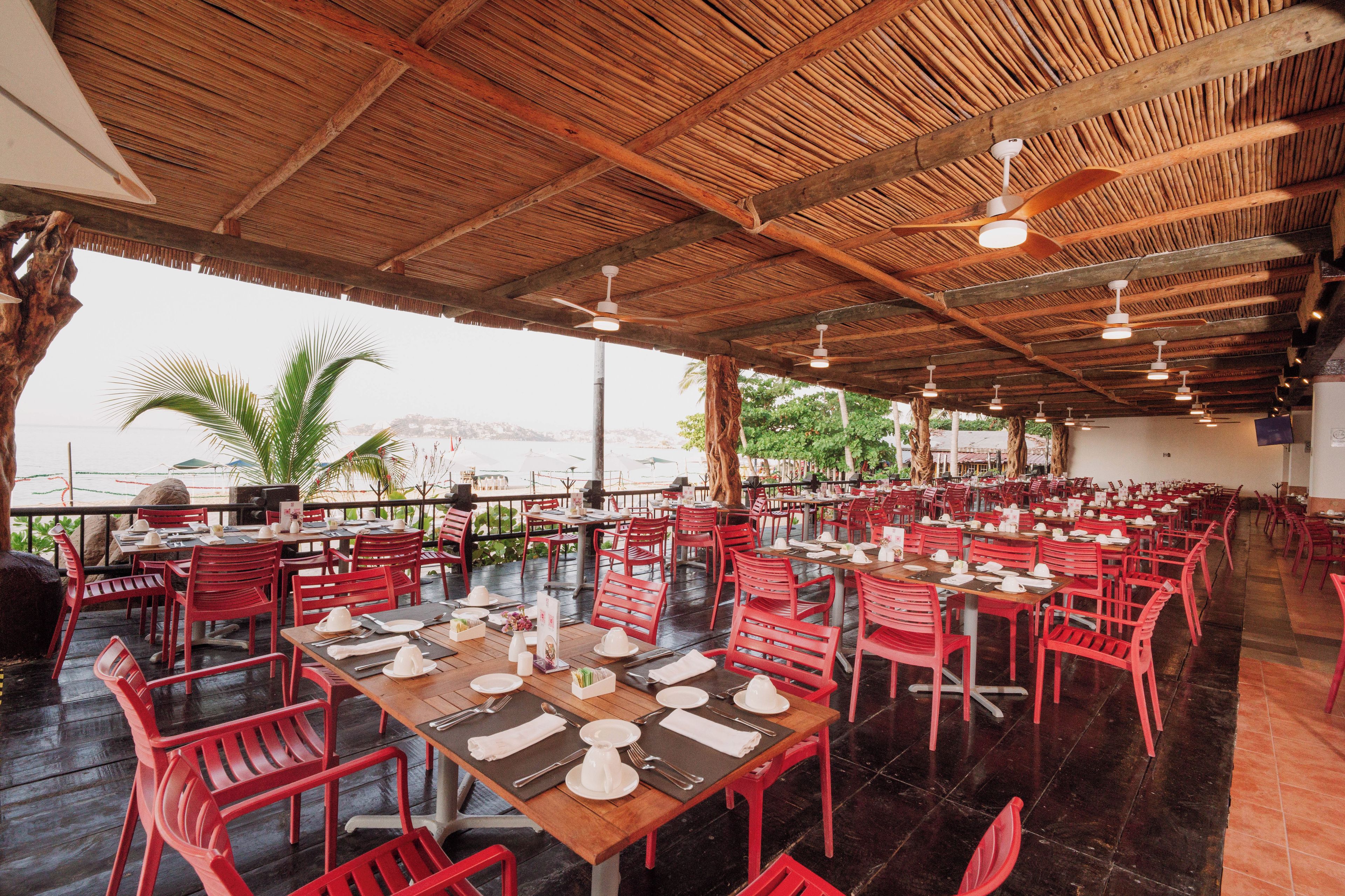 An outdoor restaurant with wooden tables and red chairs, set against a backdrop of a beach and palm trees.