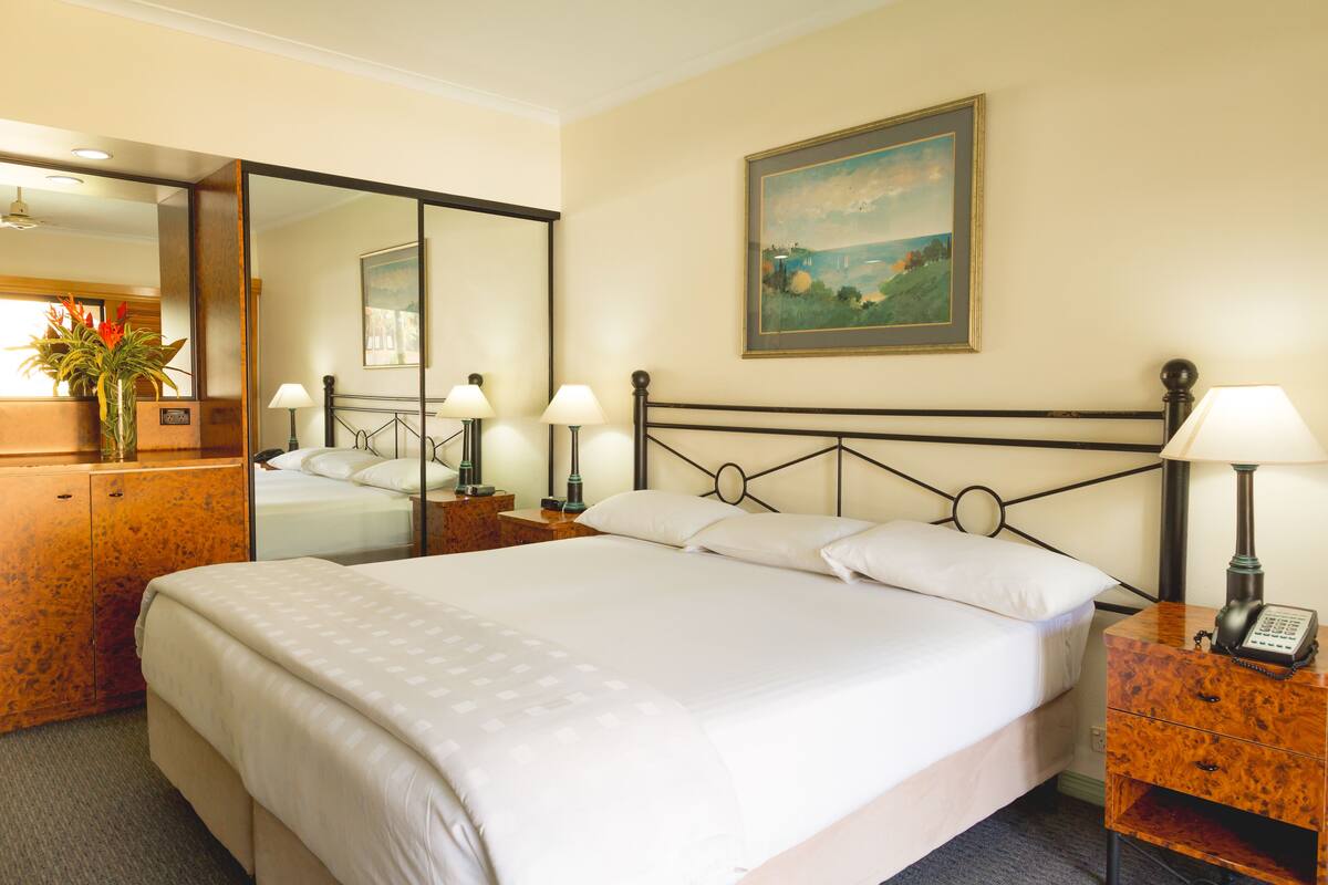 A hotel room with a large bed, a wooden nightstand with a lamp, a mirror, and a painting on the wall.