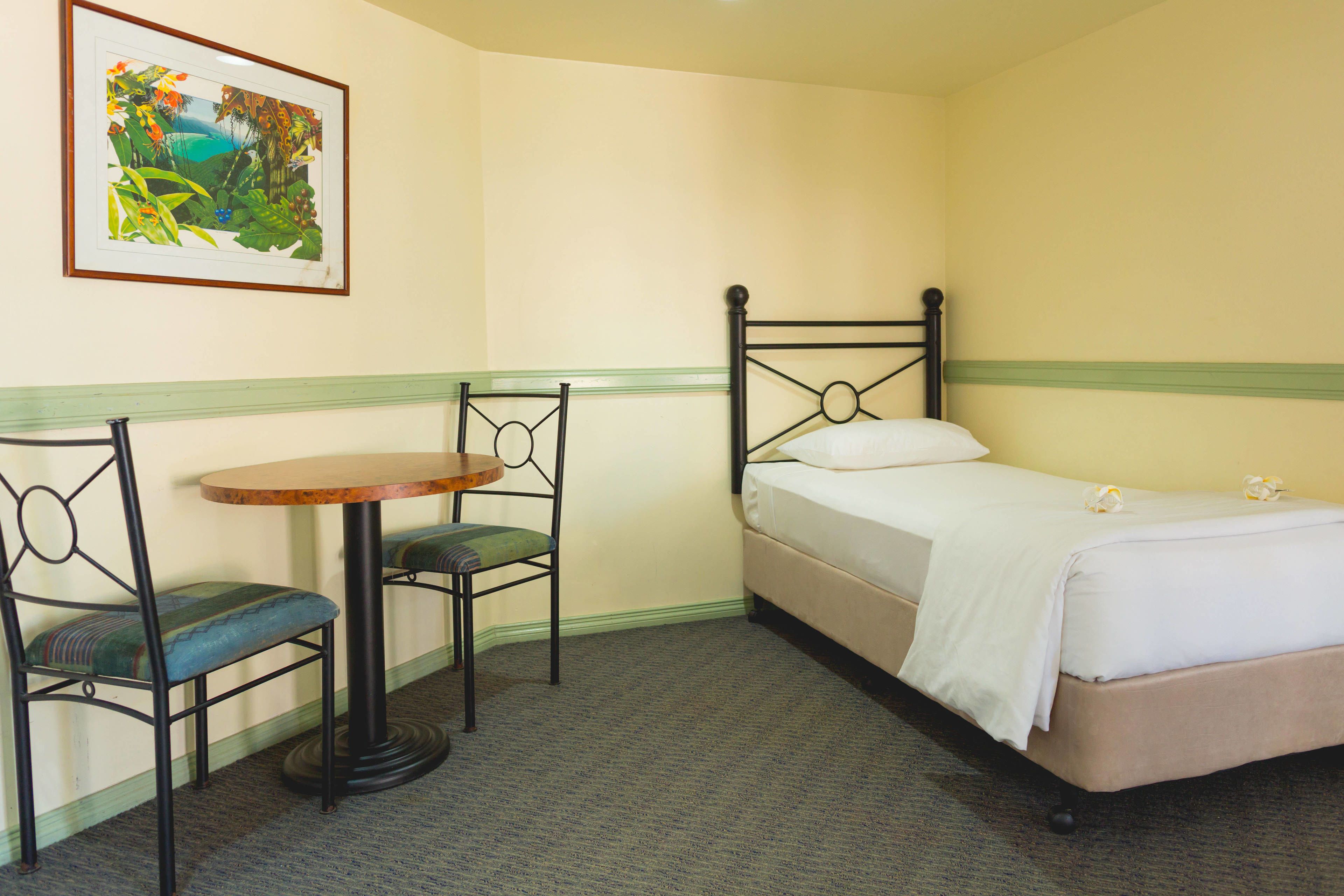 A small, neatly arranged room with a single bed, a chair, a small table, and a framed picture on the wall.