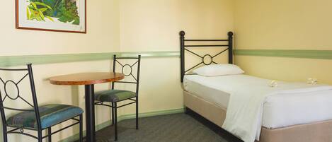 A small, neatly arranged room with a single bed, a chair, a small table, and a framed picture on the wall.