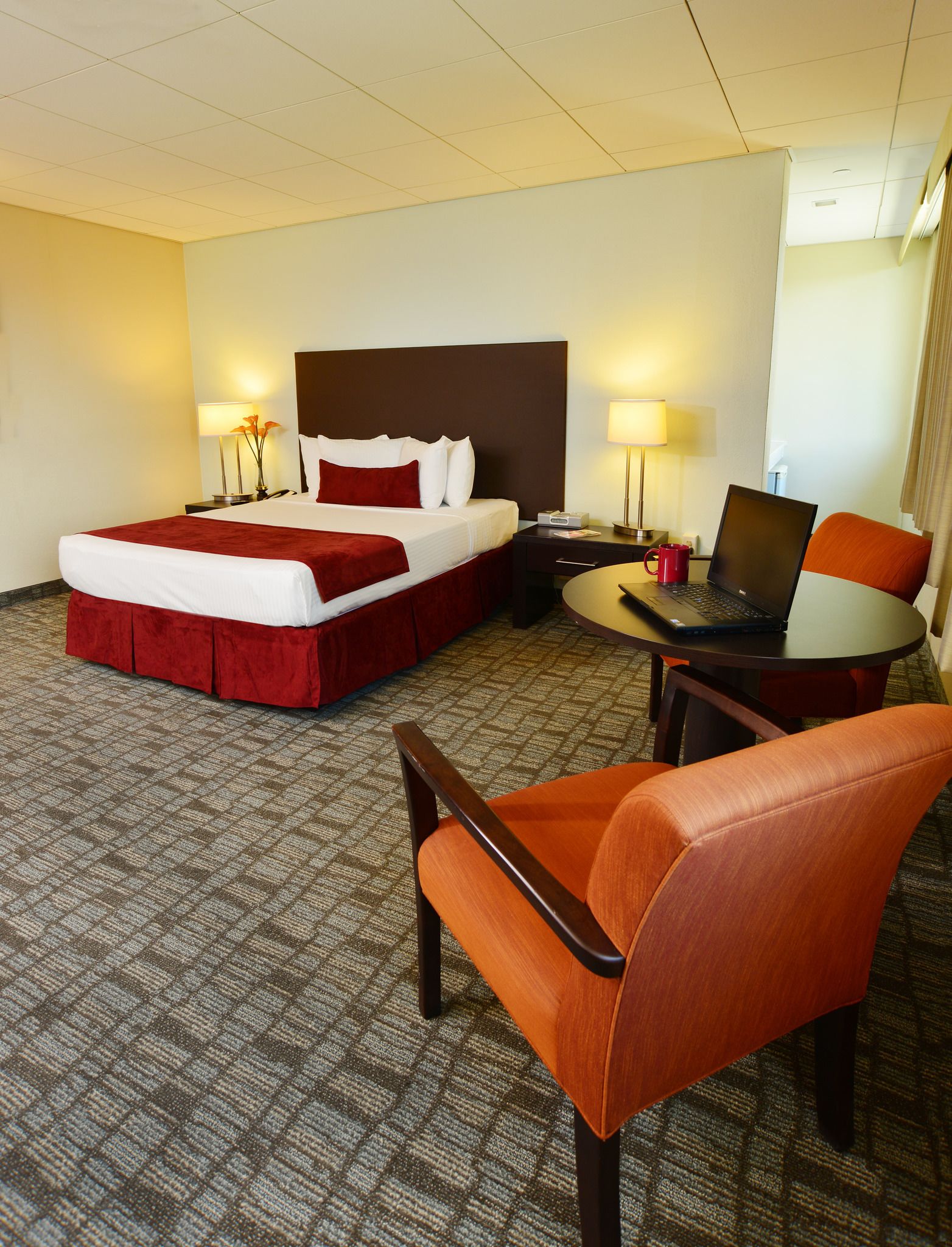 A hotel room with a bed, a desk with a laptop, a chair, and a lamp.