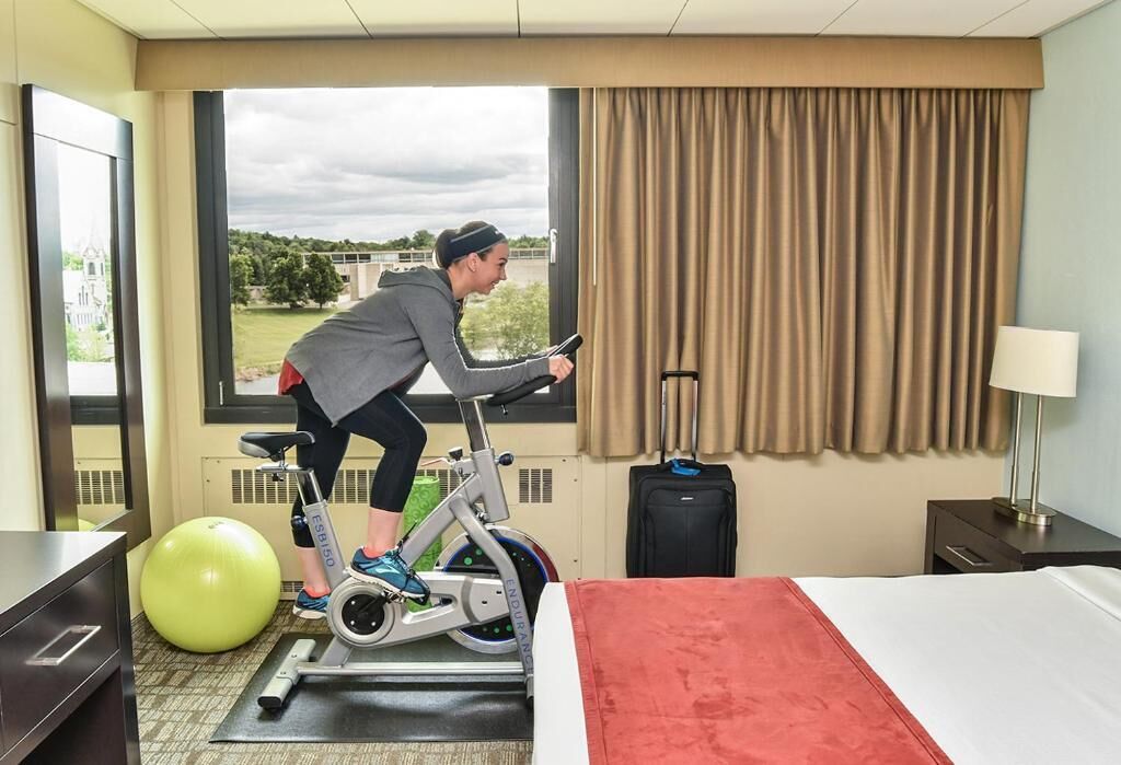 A hotel room with a bed, a treadmill, a green exercise ball, and a window with a view of a bridge and greenery.