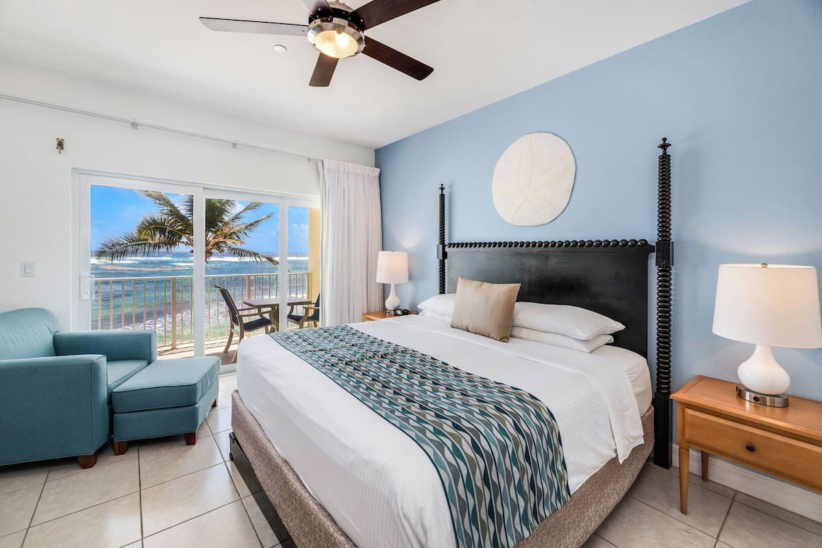 A bedroom with a bed, a ceiling fan, a view of the ocean, a chair, and a nightstand with a lamp.