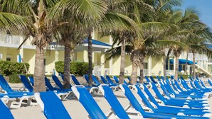 A row of blue and white sun loungers lined up on a sandy beach, with palm trees and a building in the background. - The Reef Beach Resort, East End, Grand Cayman (East End)