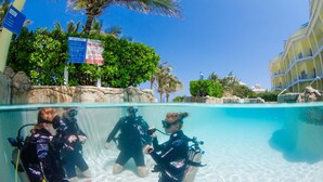A clear pool with three divers, a lifeguard sign, and a building with balconies. - The Reef Beach Resort, East End, Grand Cayman (East End)