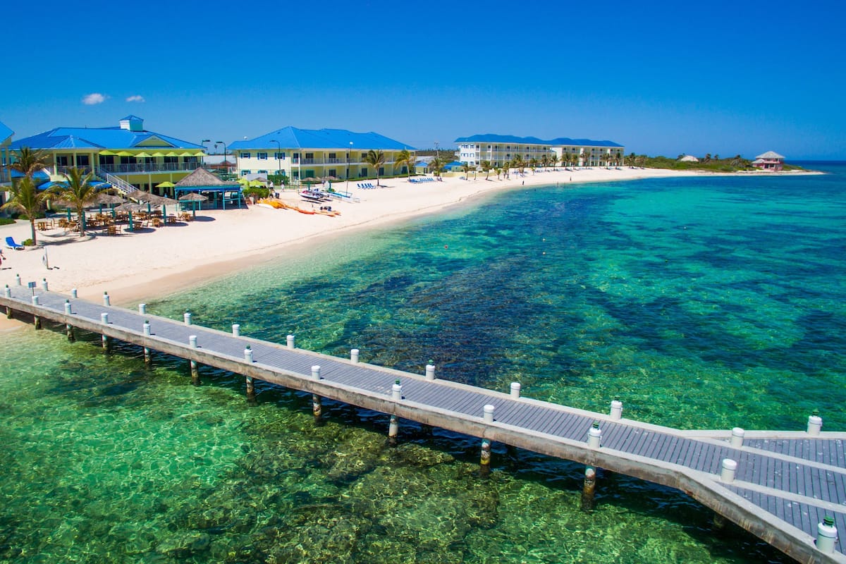 A beach resort with a pier extending into clear turquoise waters, a sandy beach, and resort buildings with multiple balconies.