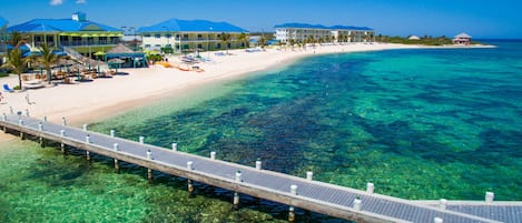 Ett semesterhotell vid stranden med en brygga som sträcker sig ut i det klara, turkosfärgade vattnet, en sandstrand och hotellbyggnader med flera balkonger.