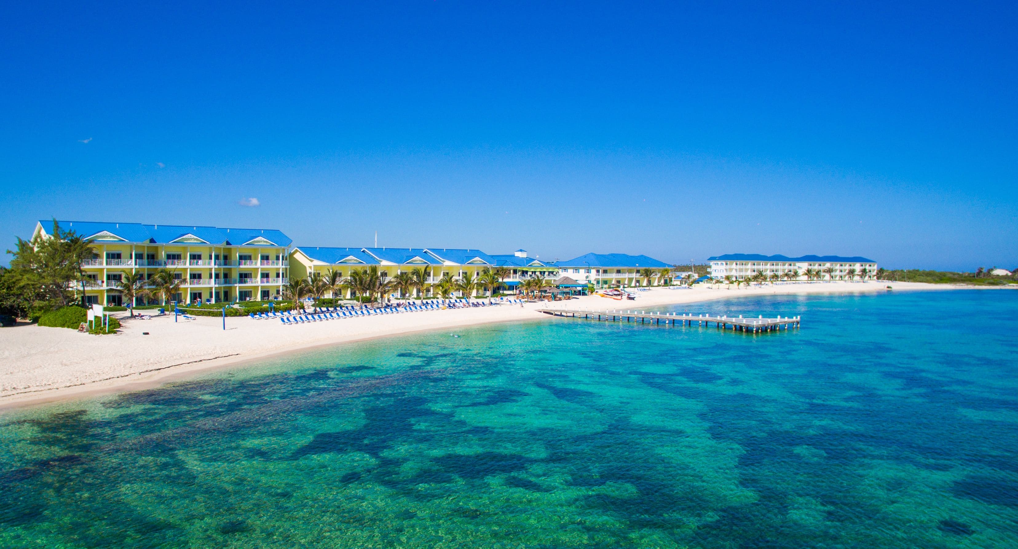 A beachfront hotel with multiple buildings, a sandy beach, clear blue water, and a pier extending into the sea.