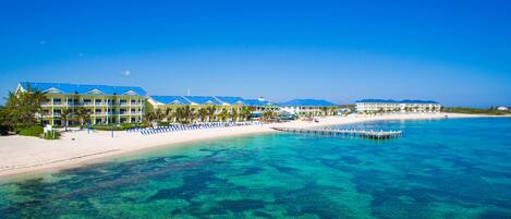 A beachfront hotel with multiple buildings, a sandy beach, clear blue water, and a pier extending into the sea.