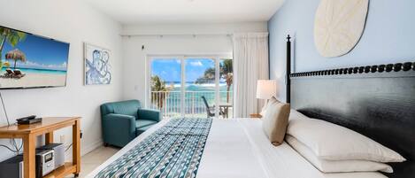 A hotel room with a bed, a ceiling fan, a TV, a desk with a computer, a chair, and a view of the ocean.