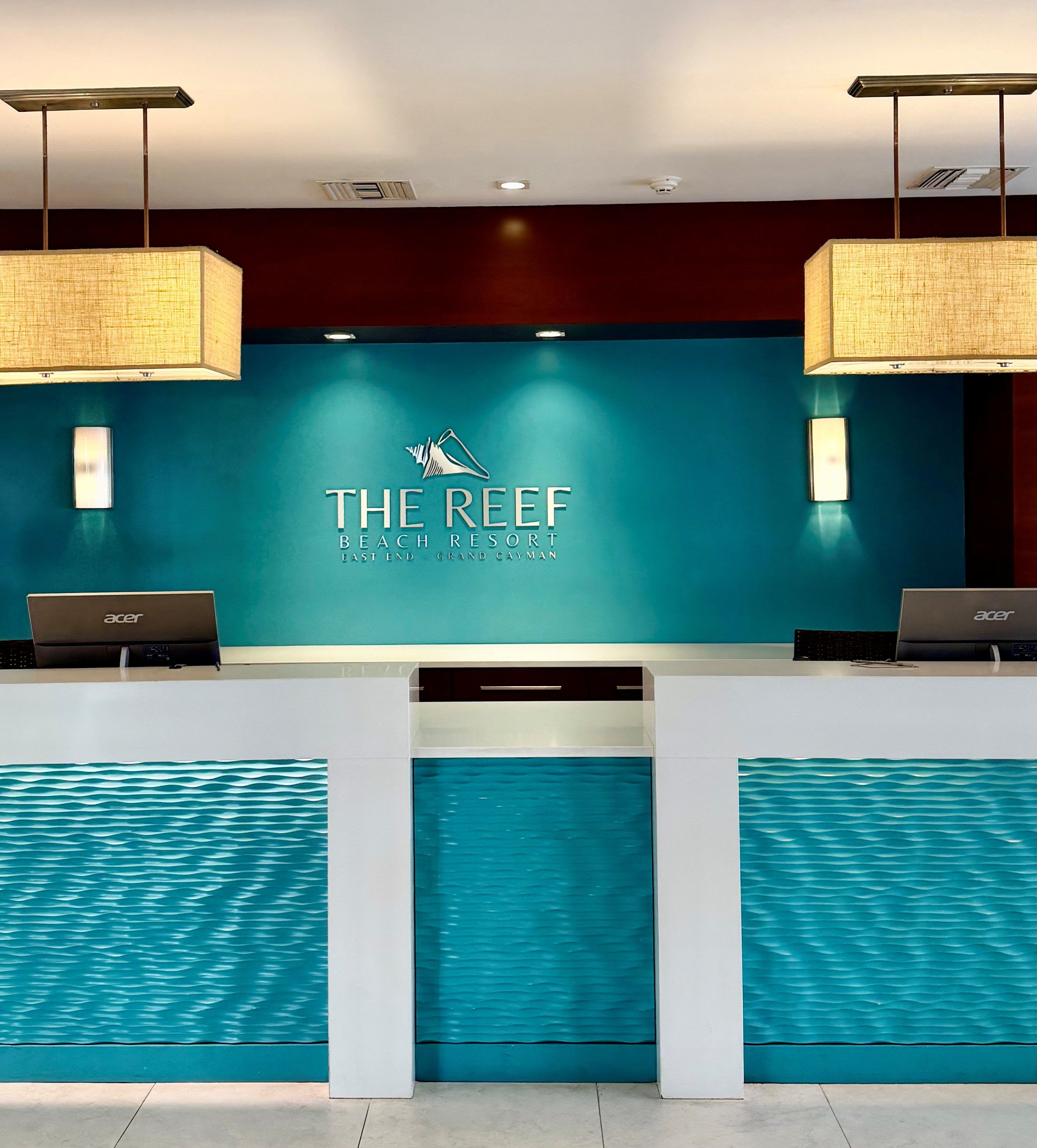 A reception desk at The Reef Beach Resort with a logo, two laptops, and a textured wall design.