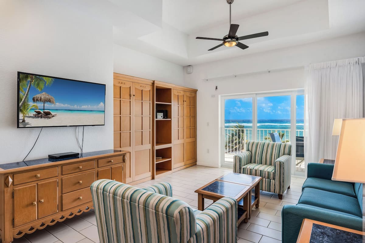A living room with a TV, striped chairs, a sofa, a ceiling fan, and a view of the ocean.