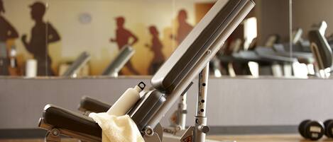 A gym with exercise equipment, including a bench and weights, and a mirror reflecting silhouettes of people exercising.