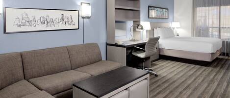 A hotel room with a sofa, a desk, a bed, and a cityscape artwork on the wall.