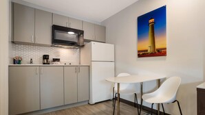 Une cuisine moderne avec des meubles gris, un réfrigérateur blanc, un four à micro-ondes et une machine à café. On y trouve une table blanche avec deux chaises. Un tableau représentant un phare est accroché au mur.