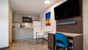 Une cuisine moderne équipée d’un four à micro-ondes, d’un réfrigérateur et de meubles blancs. On y trouve un coin repas avec une table et des chaises blanches, ainsi qu’une chaise bleue près d’un bureau. Un téléviseur à écran plat est fixé au mur.