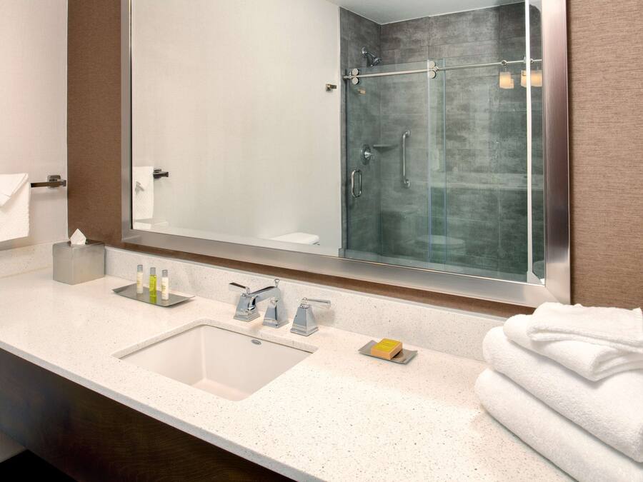 A modern bathroom with a large mirror, a glass shower enclosure, a sink with toiletries, and neatly stacked towels.