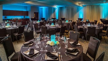 Un salón de banquetes con mesas redondas dispuestas para comer, una zona de bar y un amplio ventanal con cortinas.