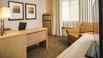 A hotel room with a wooden desk, a brown leather chair, a sofa, a standing heater, and two framed artworks on the wall.