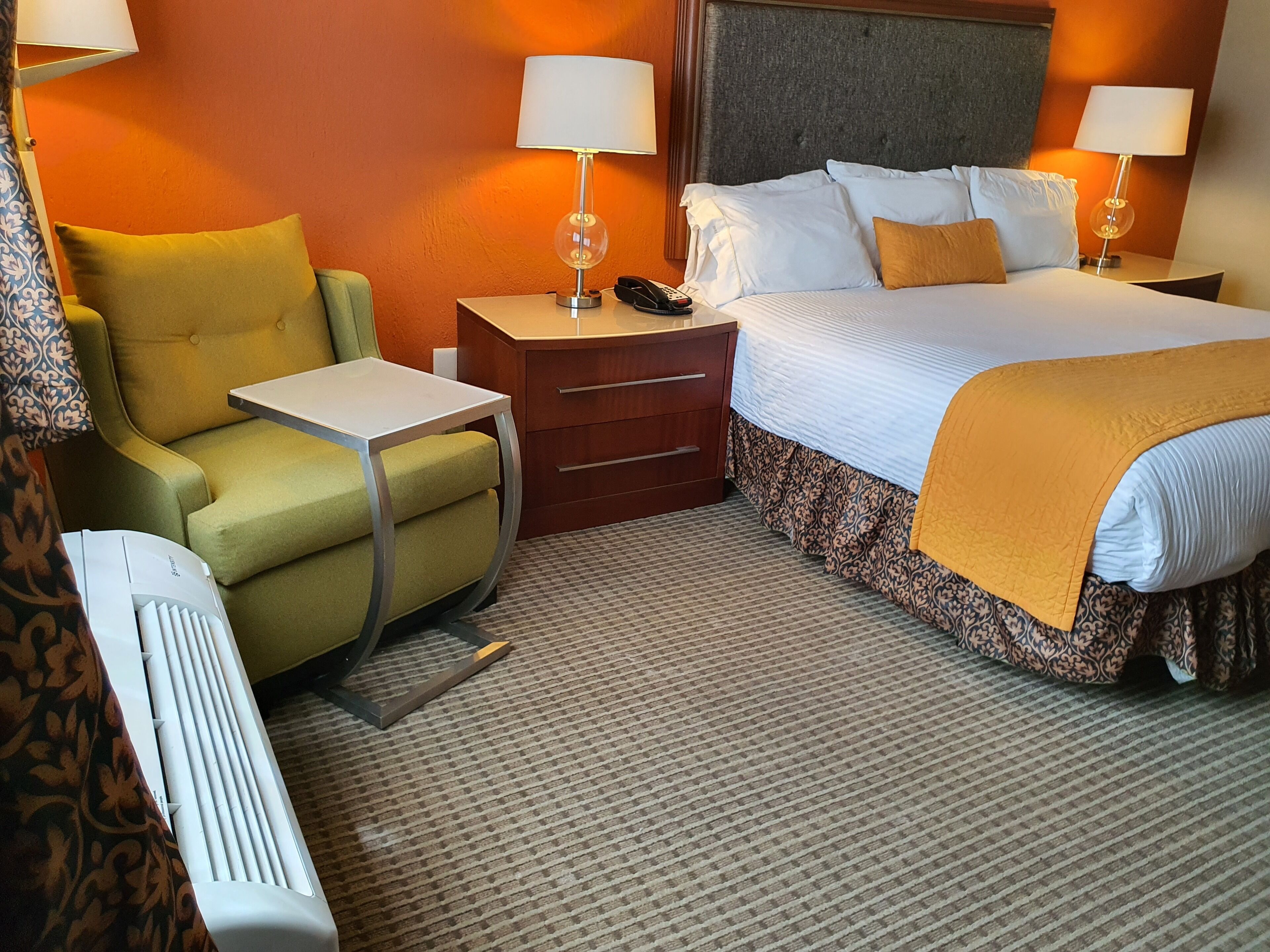 Una habitación de hotel con cama, mesita de noche, lámpara, silla y un calentador.