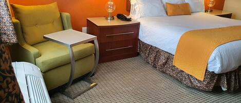 Una habitación de hotel con cama, mesita de noche, lámpara, silla y un calentador.