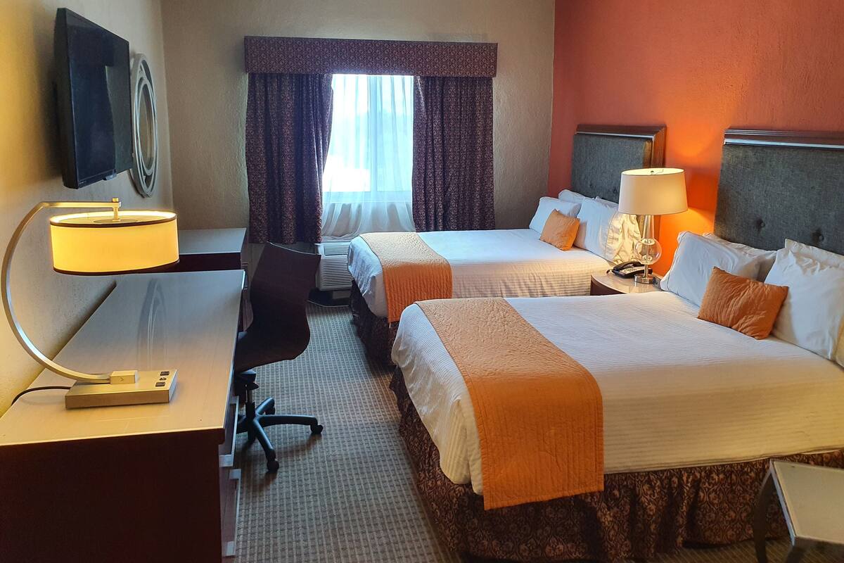 Ein Hotelzimmer mit zwei Betten, einem Schreibtisch mit Lampe, einem Sessel und einem an der Wand montierten Fernseher.