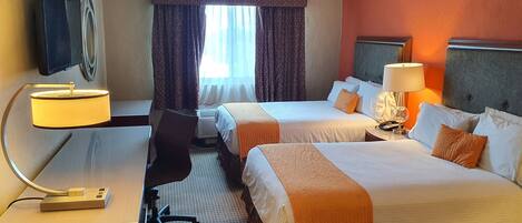 A hotel room with two beds, a desk with a lamp, a chair, and a TV mounted on the wall.