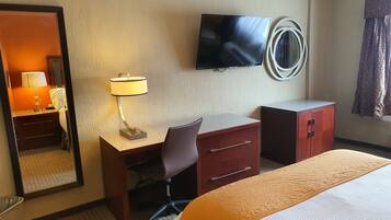 Una habitación de hotel con cama, un escritorio con silla, un televisor montado en la pared y un espejo.