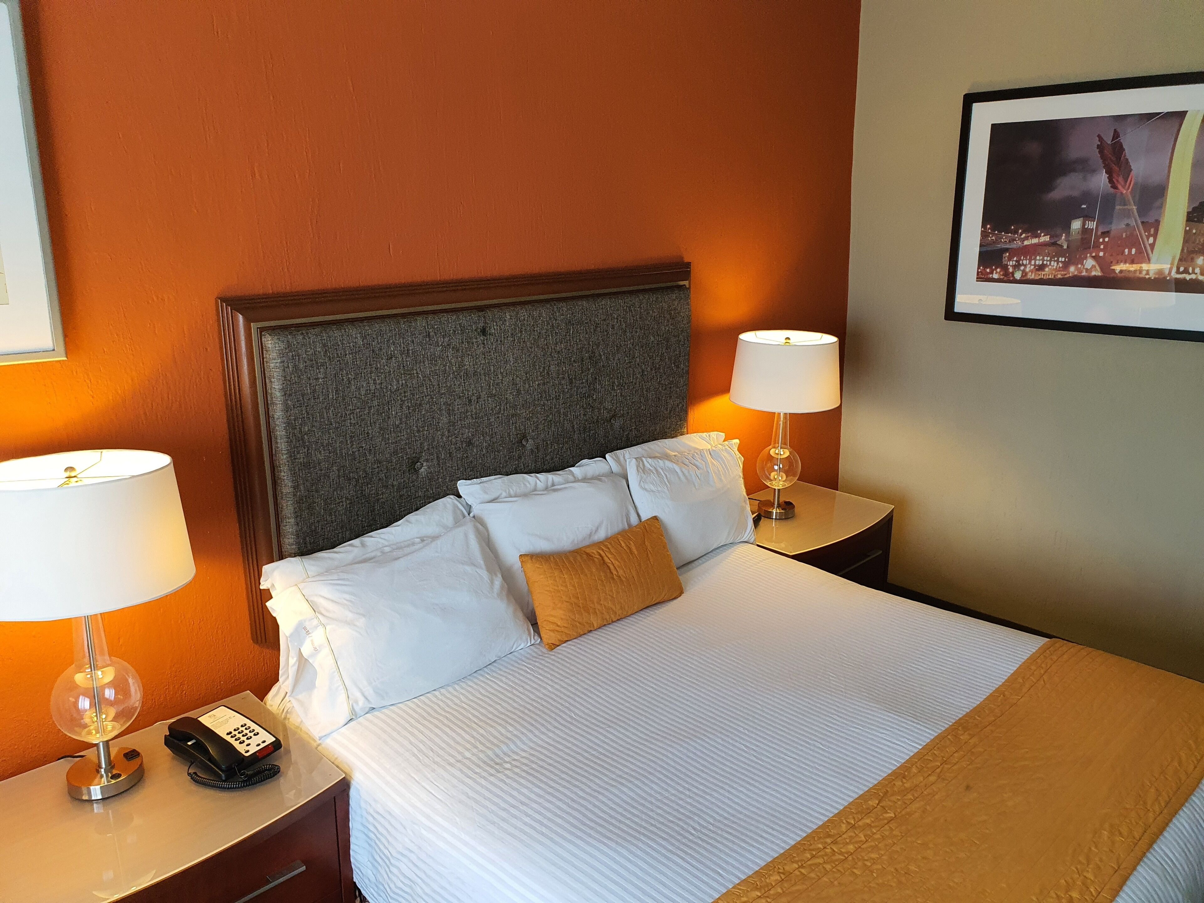 Habitación de hotel con cama, dos mesitas de noche con lámparas, teléfono y un cuadro en la pared.