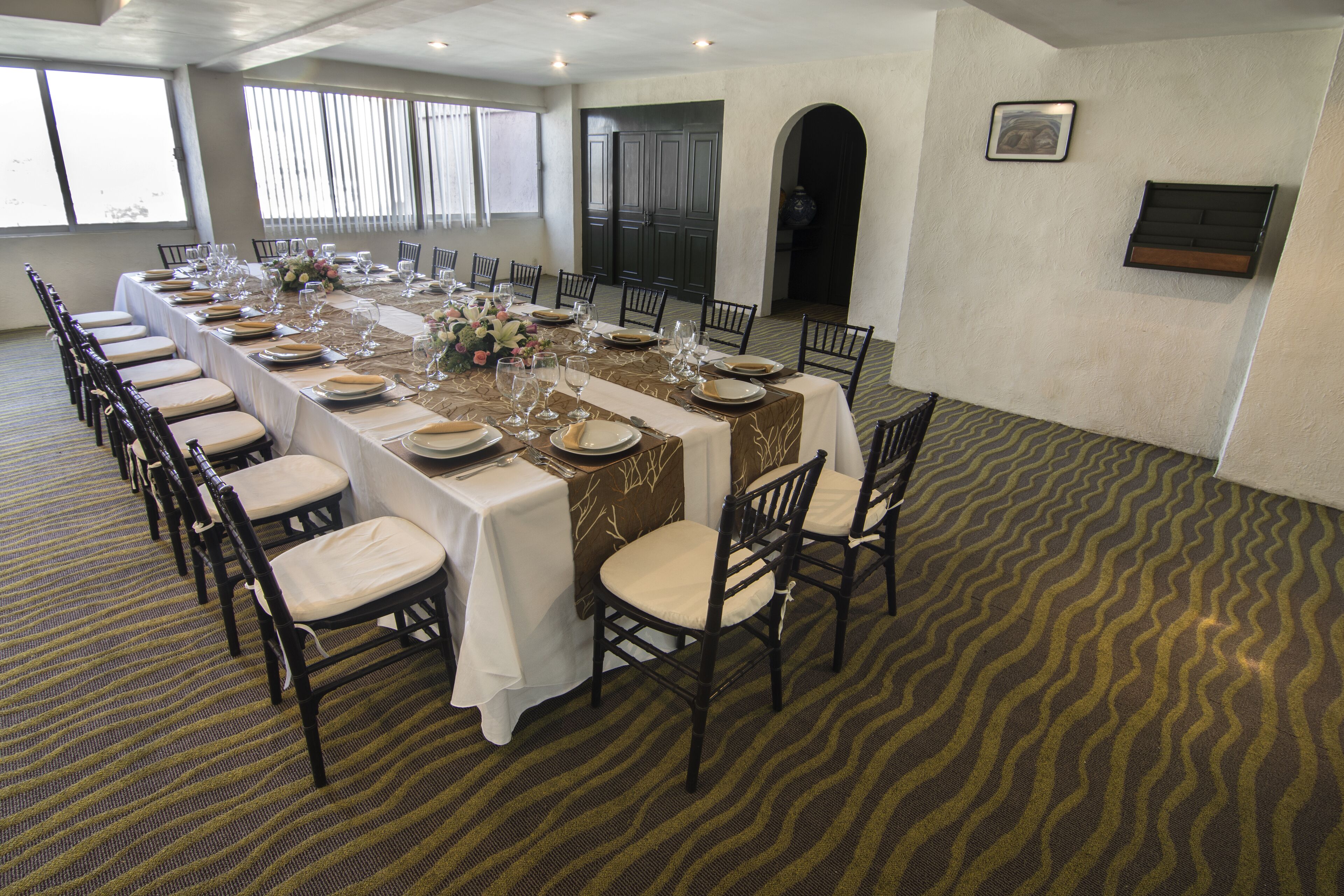 A long dining table set for a formal event with chairs, a patterned carpet, and a view of the outside through large windows.