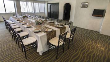 A long dining table set for a formal event with chairs, a patterned carpet, and a view of the outside through large windows.
