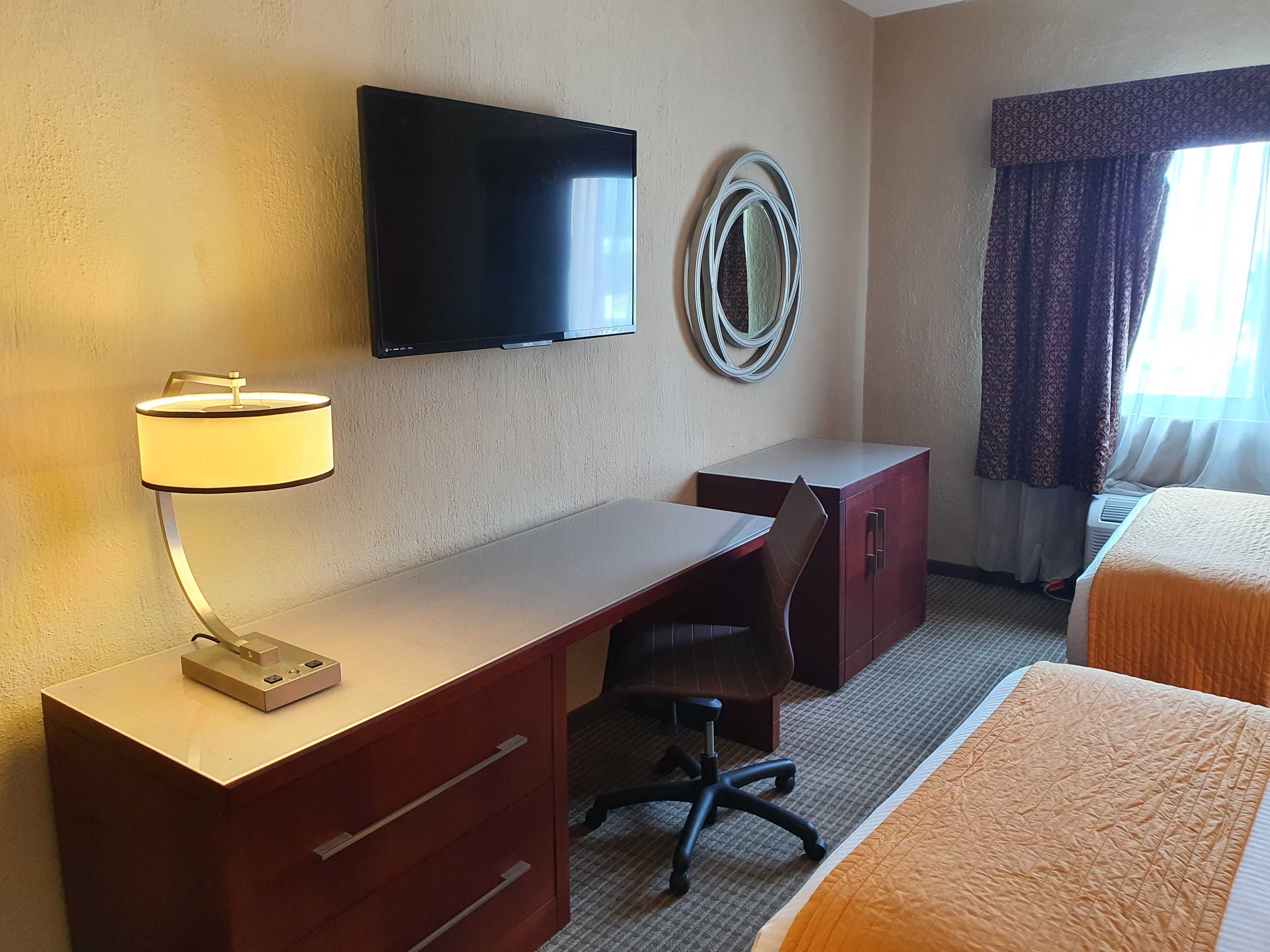 Habitación de hotel con televisión de pantalla plana en la pared, un escritorio con lámpara, una silla, una cama con colcha naranja y una ventana con cortinas estampadas.
