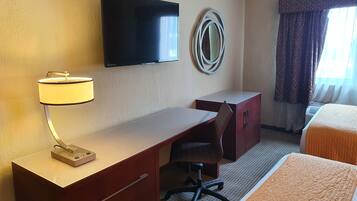Habitación de hotel con televisión de pantalla plana en la pared, un escritorio con lámpara, una silla, una cama con colcha naranja y una ventana con cortinas estampadas.