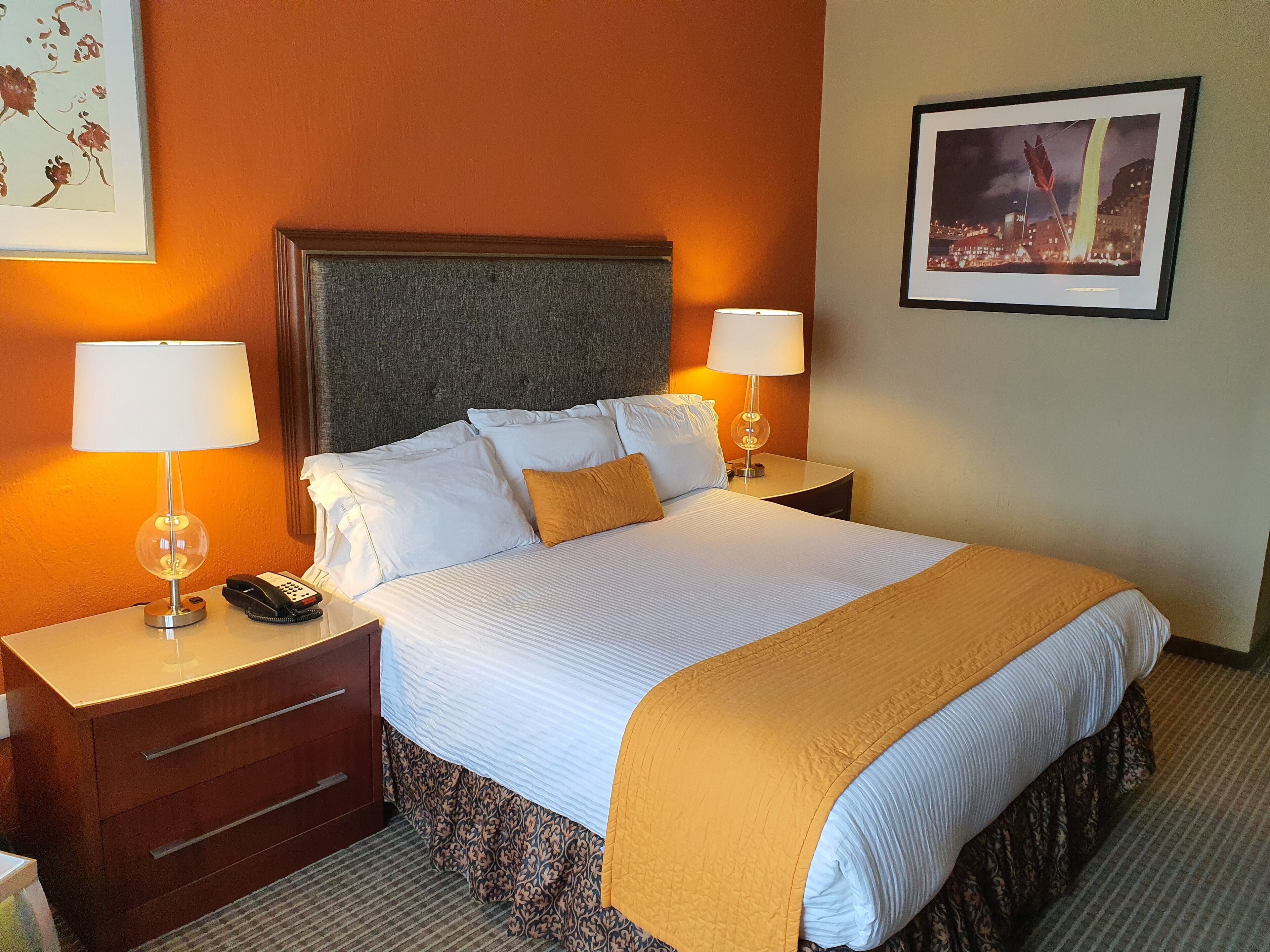 Une chambre d’hôtel avec un grand lit, deux tables de chevet avec des lampes, une petite table en bois, un couvre-lit à motifs et un tableau encadré au mur.