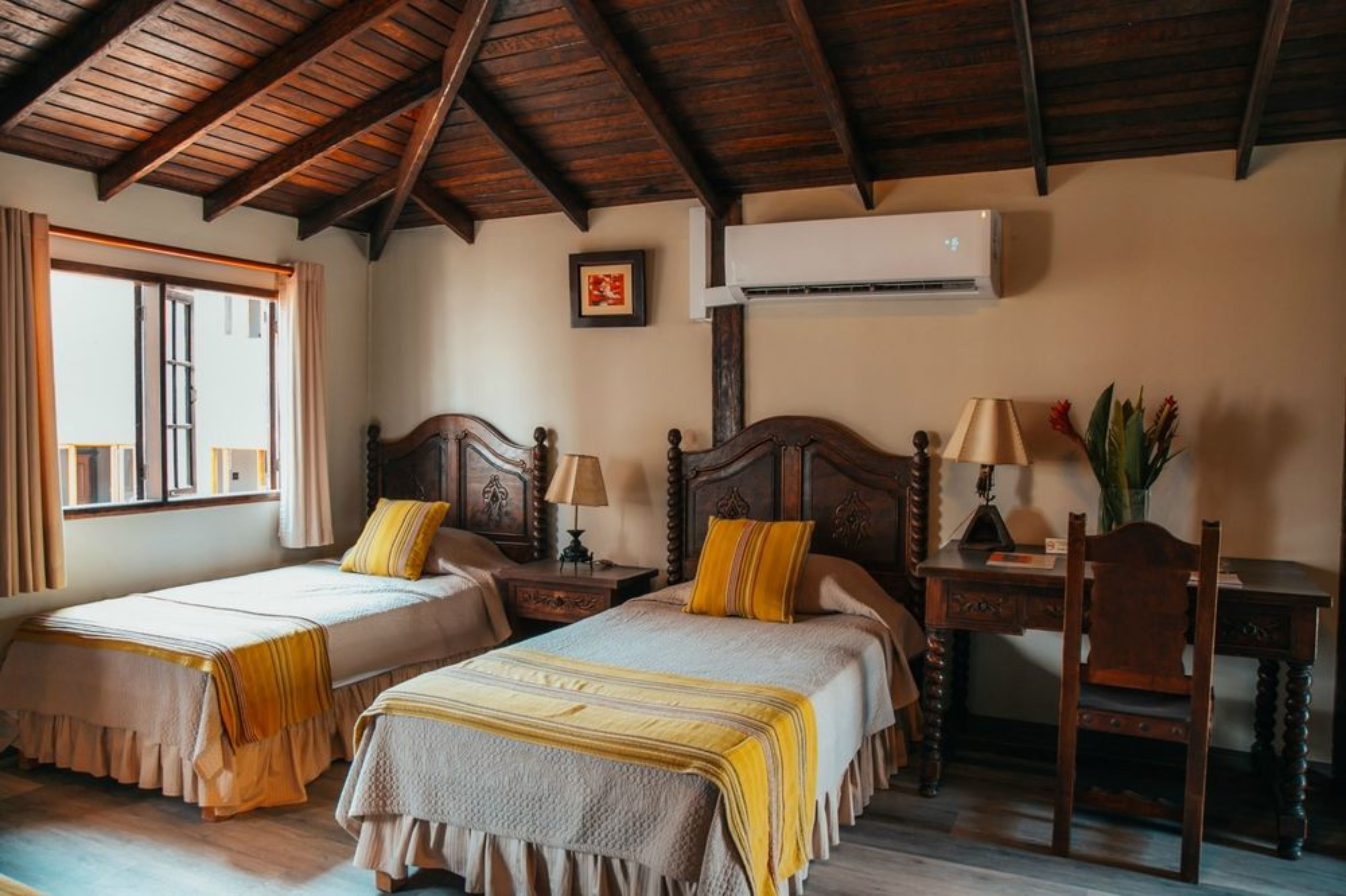 A room with two beds, wooden furniture, a desk, and an air conditioning unit.