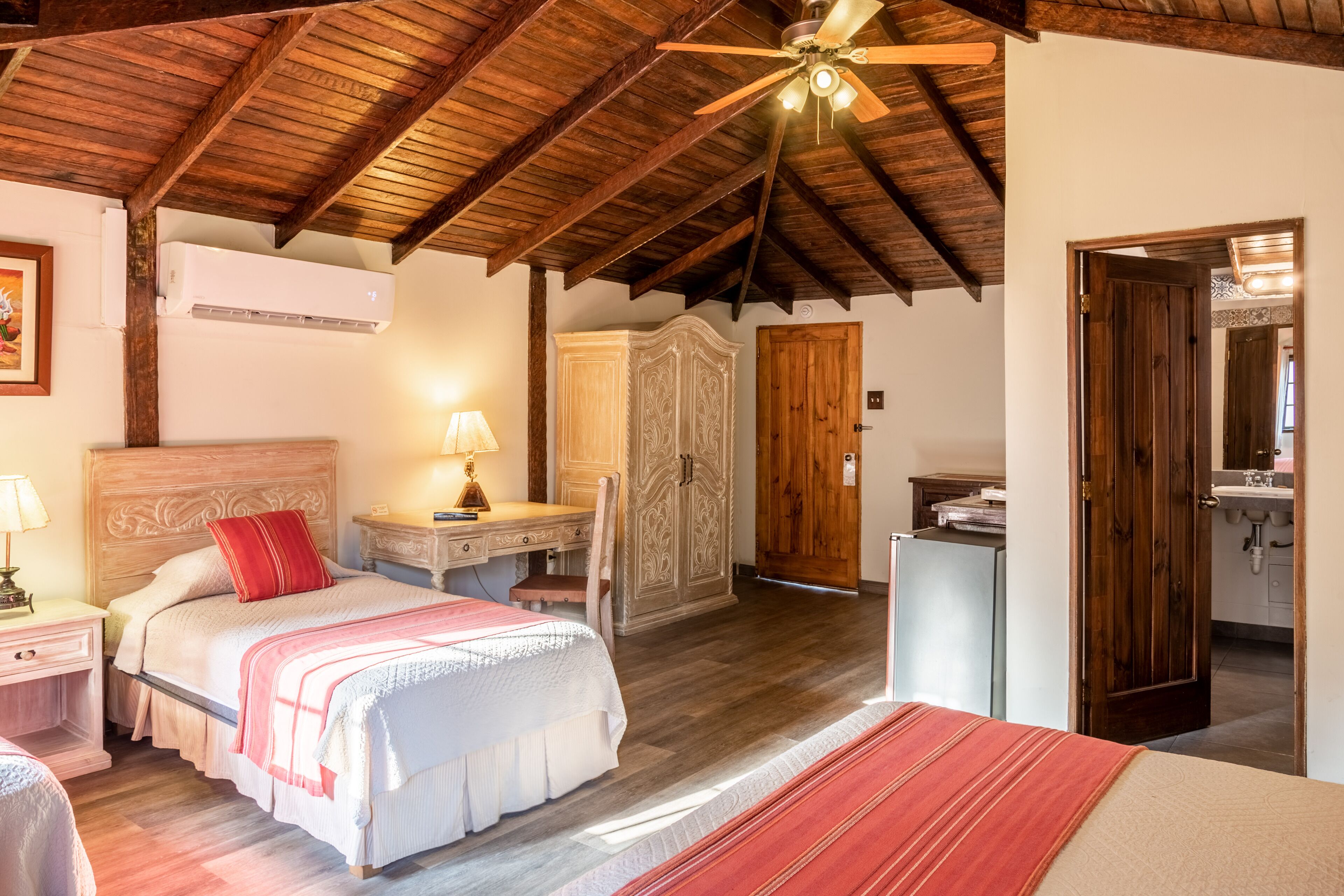A bedroom with a wooden ceiling, a bed with a red pillow, a nightstand, a desk, and a wardrobe.