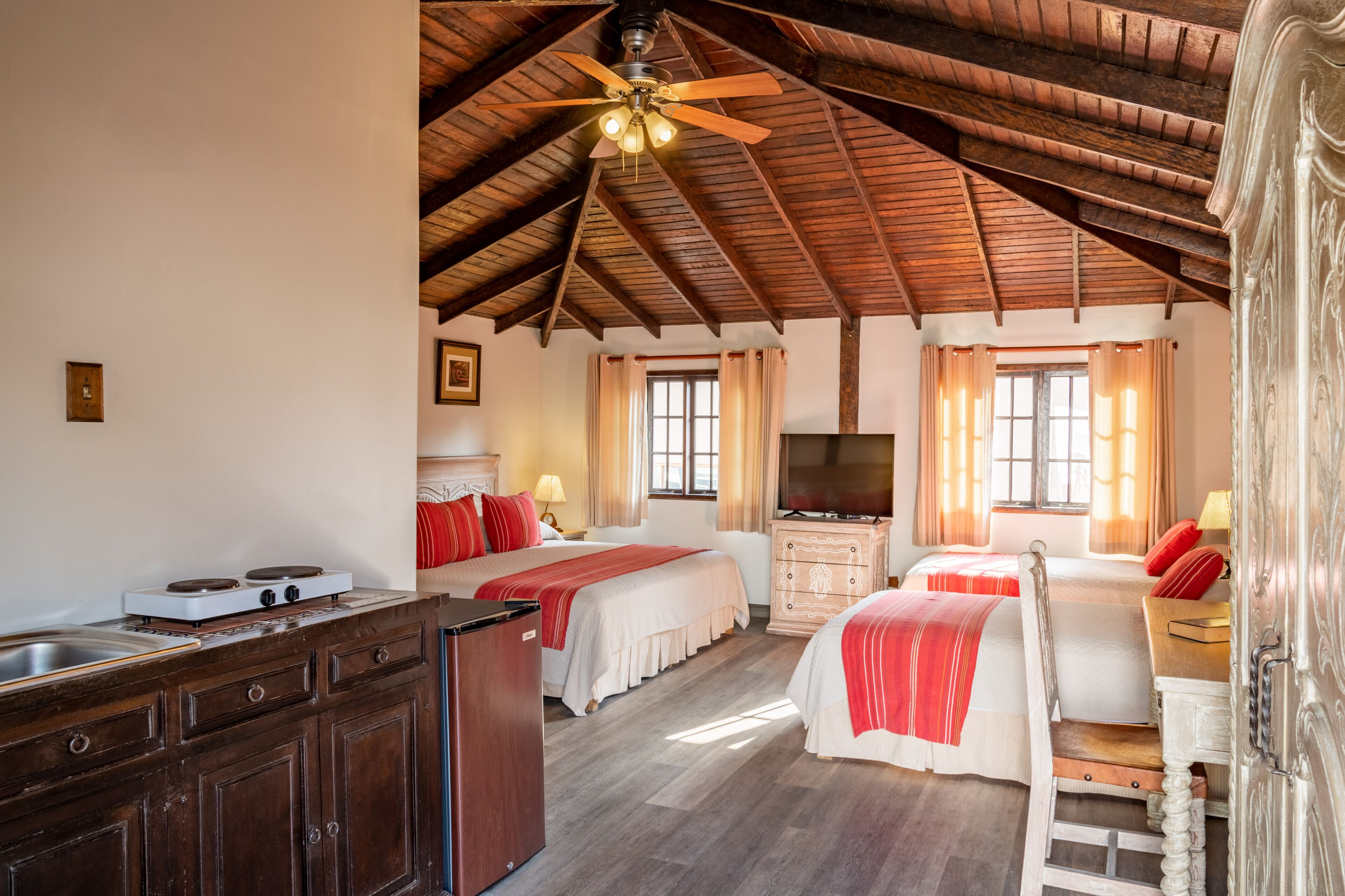Une chambre avec deux lits, une télévision, un plafond en bois et un coin cuisine équipé d’une cuisinière.