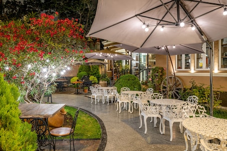 An outdoor patio with white metal chairs and tables, surrounded by greenery and red flowers.