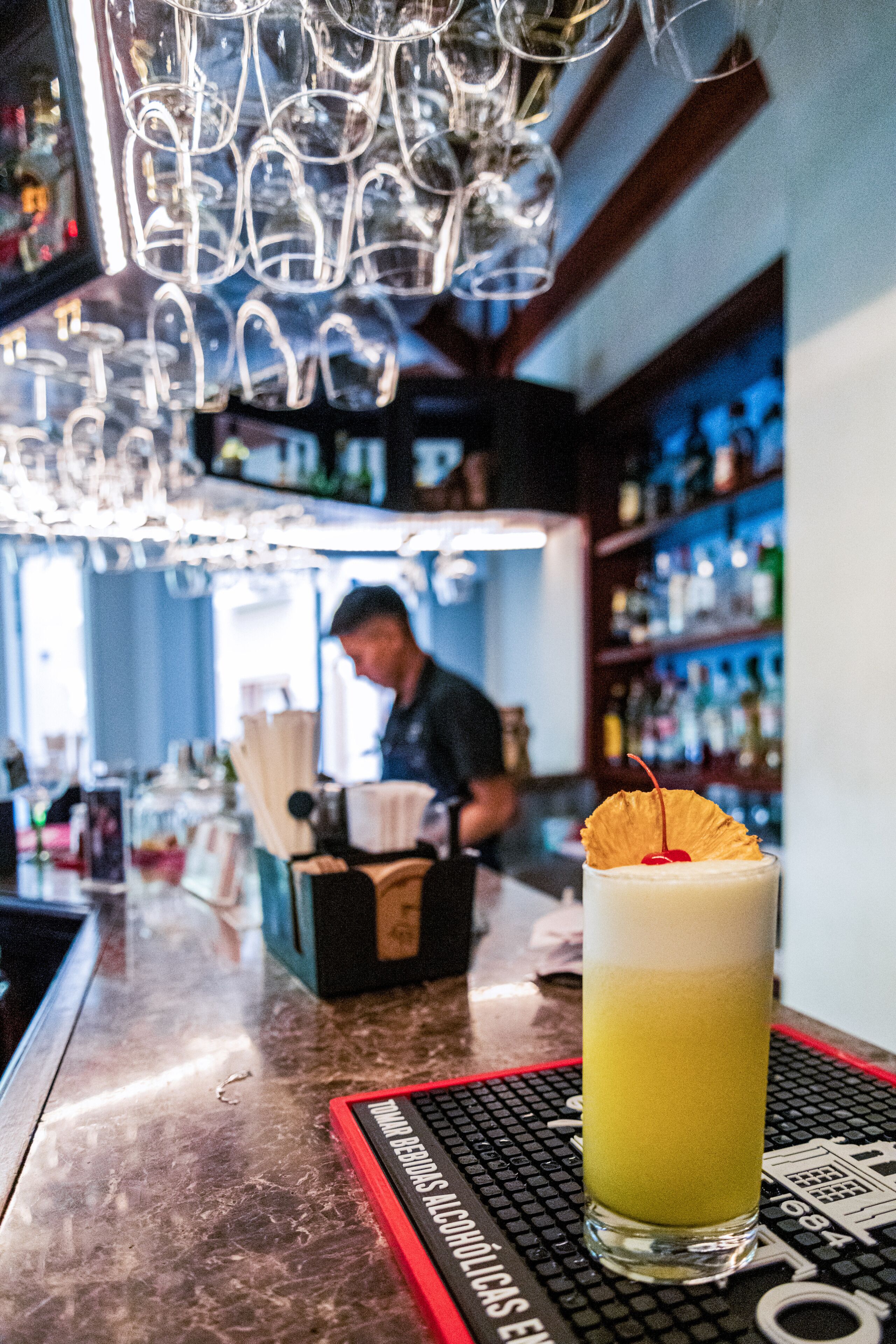 Una zona de bar con un vaso alto de una bebida amarilla, un barman al fondo y estantes llenos de botellas.