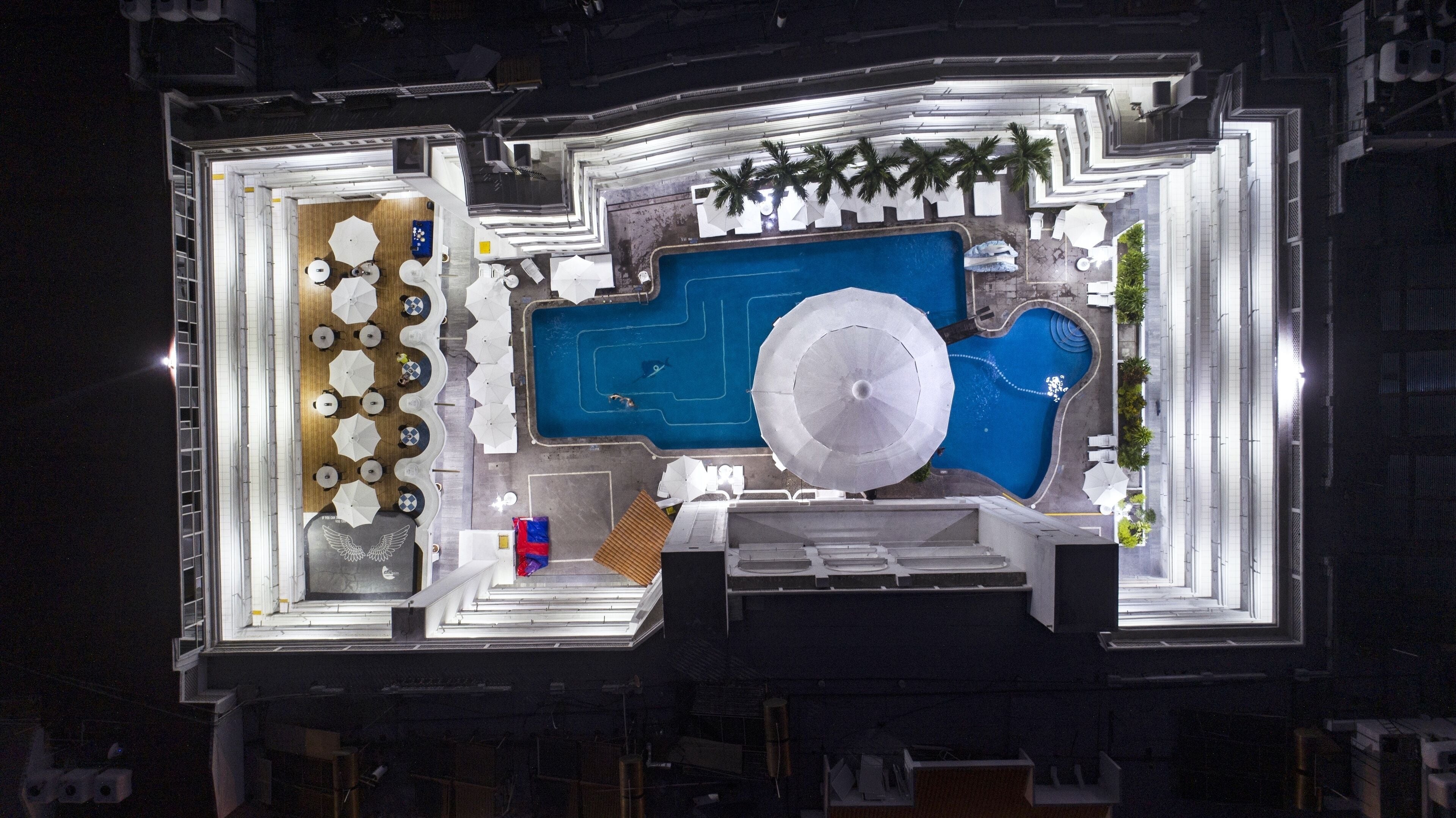 An aerial view of a hotel with a swimming pool, sun loungers, umbrellas, and a covered seating area.