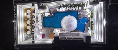 An aerial view of a hotel with a swimming pool, sun loungers, umbrellas, and a covered seating area.