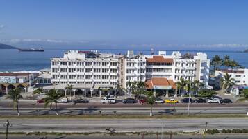 A multi-story hotel with a central courtyard, located near a coastal road with palm trees and a view of the sea.