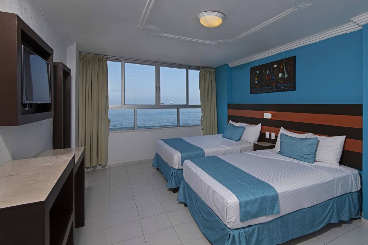 A hotel room with two beds, a TV, a window with a view of the sea, and a painting on the wall.